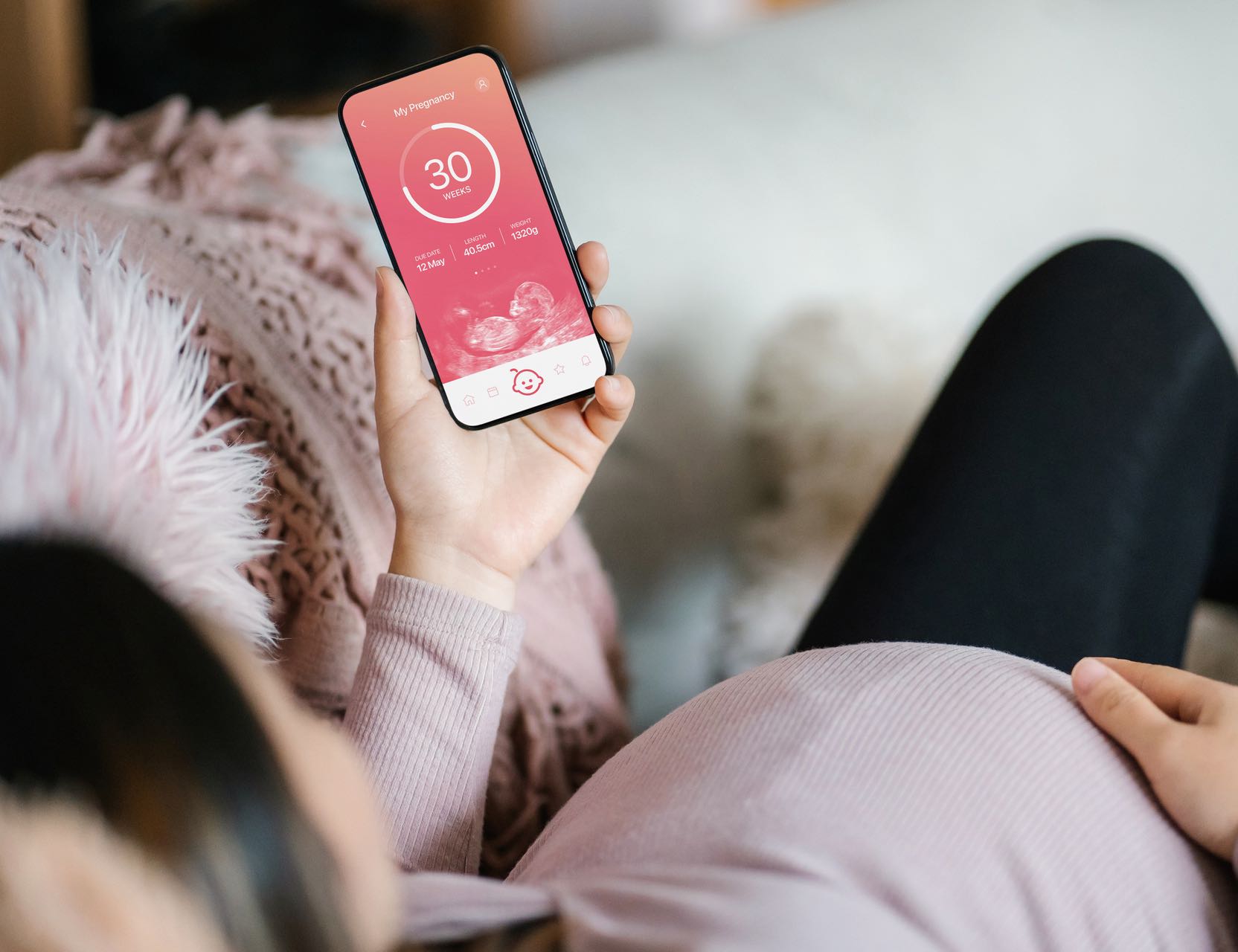 Young Asian pregnant woman lying on sofa in the living room at cozy home, using baby tracker app on smartphone to monitor her baby's growth and development throughout the pregnancy journey. Technology and pregnancy lifestyle